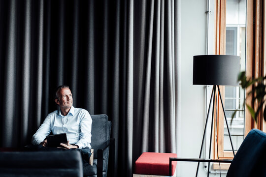 Thoughtful male entrepreneur with digital tablet looking away while sitting on armchair in cafeteria at office