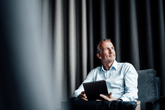 Contemplating businessman with digital tablet looking away while sitting on armchair in office cafeteria - Powered by Adobe