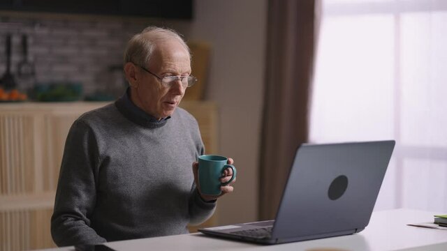 grandfather is communicating by video call, sitting alone at home kitchen, waving hands to web camera of laptop, welcome interlocutors