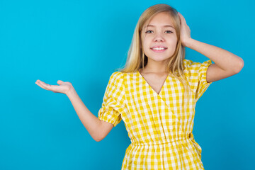 Fototapeta premium Shocked amazed surprised beautiful Caucasian little girl wearing yellow dress over blue background hold hand offering proposition