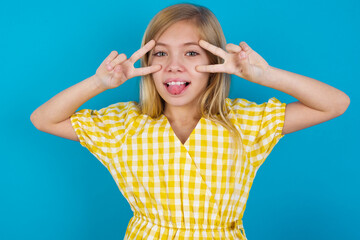 Cheerful positive beautiful Caucasian little girl wearing yellow dress over blue background shows v-sign near eyes open mouth