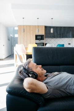 Man with hands behind head listening music through headphones while relaxing on sofa at home