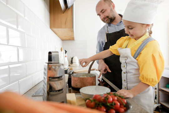 Smiling daughter wearing apron and chef's hat cooking food while standing by father in kitchen
