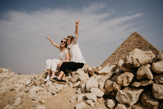 Egypt, Cairo, Two female tourists sitting together on rocks with Great Pyramid of Giza in background - Powered by Adobe