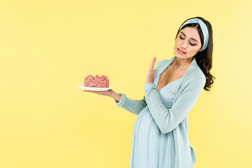pregnant woman showing refuse gesture while holding sweet dessert isolated on yellow.