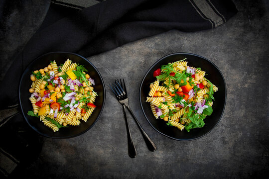 Studio shot of two bowls of fusilli pasta salad with chick-peas, bell peppers, arugula, Spanish onion, basil and parsley