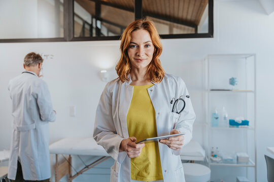 Smiling female doctor holding digital tablet while standing with coworker in background at clinic