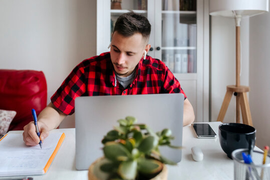 Handsome male entrepreneur with in-ear headphones writing while working on laptop at home office