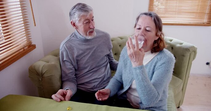 a middle-aged woman takes a pill from her husband's hand, swallows it and drinks it down with water while sitting on the couch in her house.