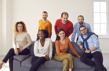 Group portrait of happy confident businessmen and businesswomen on couch in new company office. Diverse team of business partners and colleagues sitting or standing by gray sofa and smiling at camera - Powered by Adobe