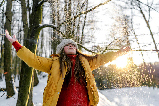 Girl in warm clothing standing with eyes closed under tree during snowfall