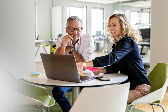 Smiling businesswoman pointing at laptop while sitting by mature businessman at table during meeting in office