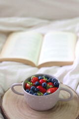 Bowl with blueberries and strawberries and open book on a bed. Selective focus. 
