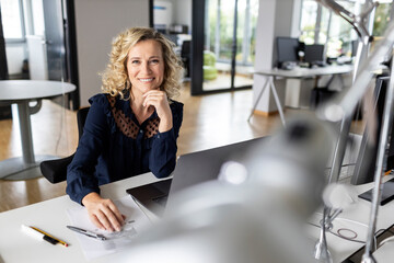Beautiful smiling businesswoman with hand on chin sitting at desk in office