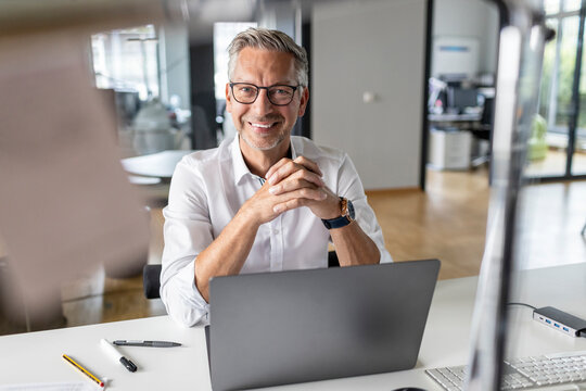 Smiling Male Entrepreneur With Laptop Sitting At Desk In Open Plan Office