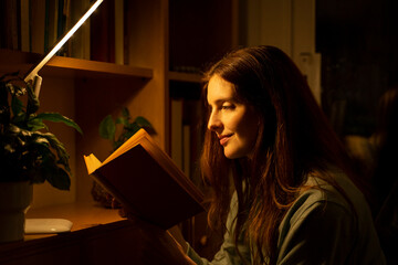 Mid adult woman reading book while sitting under illuminated lamp at home