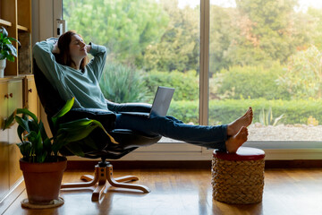 Woman with hands behind head relaxing while sitting with laptop on chair at home