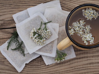 Medicinal herb Achillea millefolium with white flowers, herbal tea a mug and filter tea bags on a wooden table, top view. Healing yarrow plant for use in alternative medicine and cosmetology