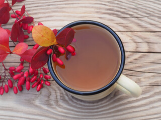 Herbal tea in an enamel mug, berberis berries and leaves  on a wooden background, top view. Healthy vitamin drink with leaves of the barberry plant and seasonal fruits to strengthen the immune system