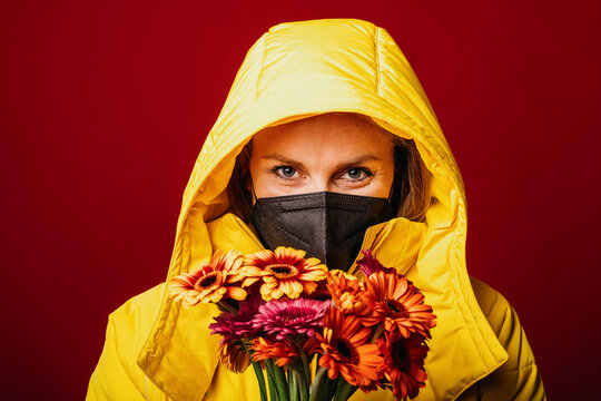 Mid adult woman wearing hood clothing and protective face mask staring while standing with bunch of flowers against red background