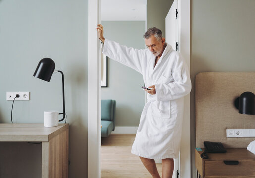 Man in bathrobe using smart phone while standing at doorway of hotel room