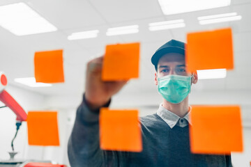 Young male engineer with protective face mask writing on adhesive notes sticked on glass wall