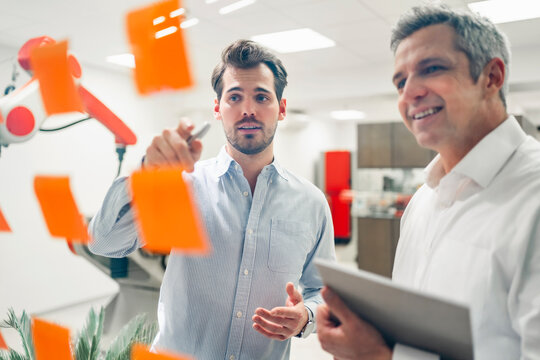 Young male engineer explaining business plans to smiling mature man in front of adhesive notes on glass wall in factory