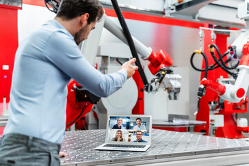 Young male automation engineer gesturing while discussing with colleagues during video call through laptop in factory