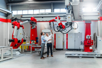 Male engineers discussing over robotic welding torch while standing at machinery in factory
