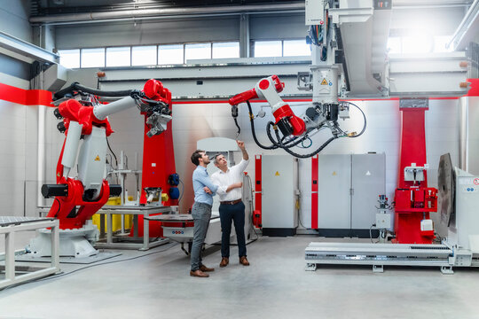 Male engineers discussing over robotic welding torch while standing at machinery in factory