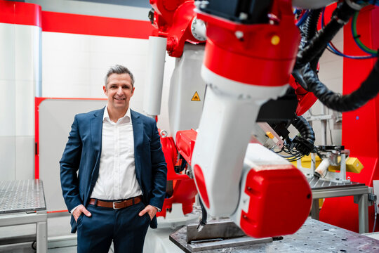 Smiling male entrepreneur with hands in pockets standing at robotics in factory