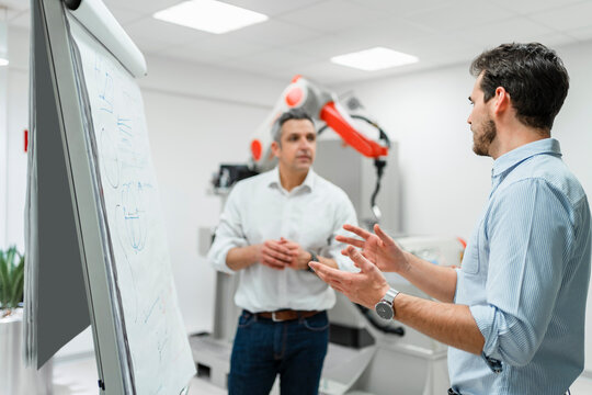 Young male engineer explaining business plans to mature entrepreneur at white board in factory