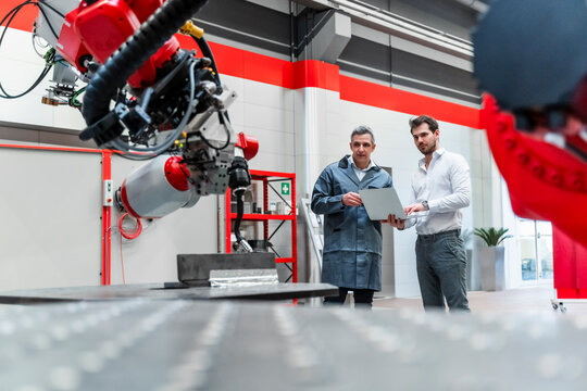 Male engineers with laptop looking at robotics while standing in factory