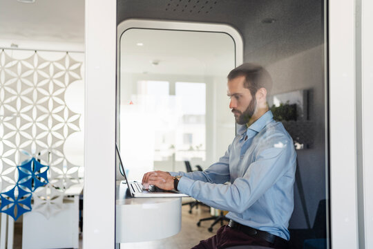 Male professional working on laptop while sitting in telephone booth at office