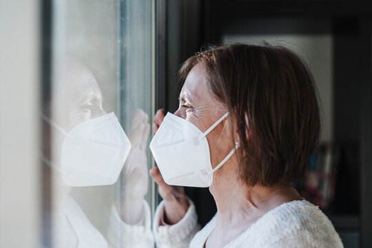 Senior woman with protective face mask looking through window during pandemic
