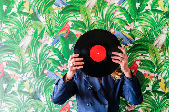 Woman covering face with record against wallpaper in jungle and parrots pattern