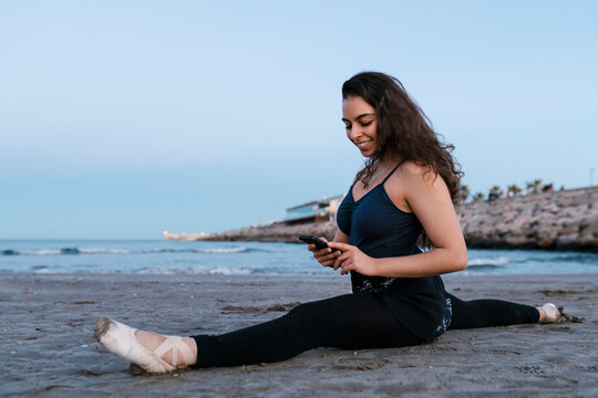 Flexible female dancer using mobile phone while doing splits at beach against clear sky