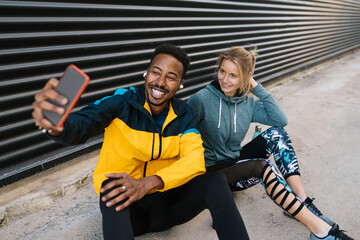 Cheerful sportsman with sportswoman taking selfie through mobile phone while sitting against wall