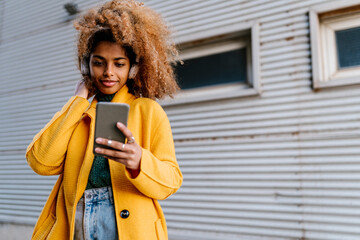 Smiling Afro woman with hand in hair listening music while using mobile phone against wall