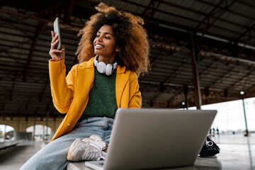 smiling Afro woman using mobile phone while sitting in front of laptop