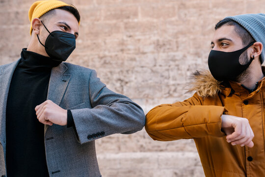 Male friends wearing protective face mask and knit hat giving elbow bump while standing against wall