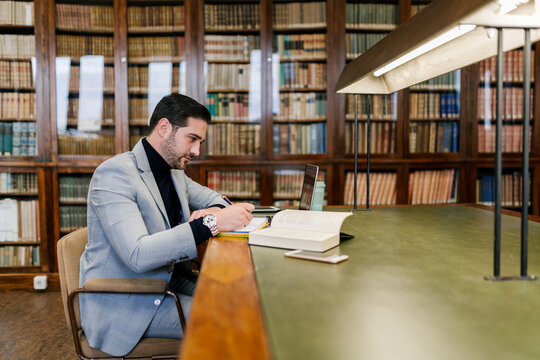 Businessman with laptop writing in book while sitting in library