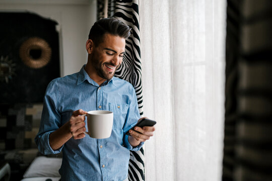 Smiling mid adult man with coffee cup using mobile phone while standing at home