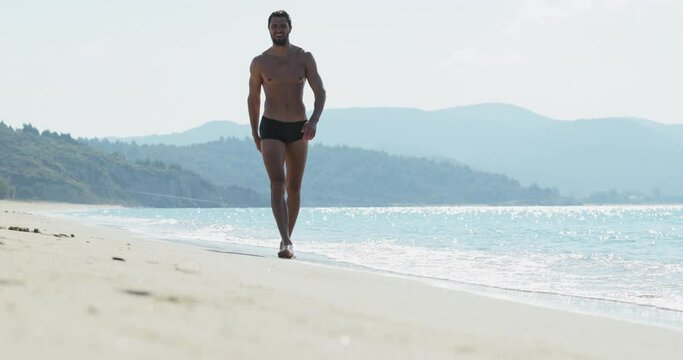 The handsome man with a perfect athletic body in swimming trunks having fun on a deserted beach in the morning, He slowly goes as the macho, showing off his body and posing