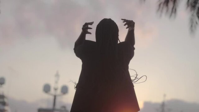 Anonymous gorgeous black woman touching long braids near palm in twilight