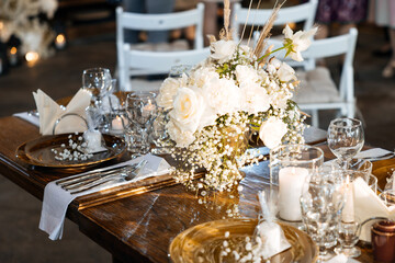 Wedding decor of roses, cotton and gypsophila and candles on a laid festive table with glasses and plates with cards. Off-season rustic wedding dinner. Selected focus and blurred background.