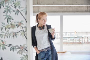 Woman with hand in pocket holding coffee cup while leaning on wall at home