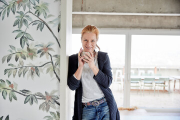 Smiling woman holding coffee cup while leaning on wall at home