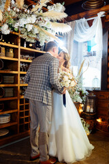 Wedding ceremony of the groom in a tuxedo and the bride in a white dress. Couple looks at each other and kisses against the background of a wedding arch indoors with dried flowers