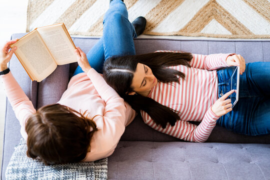 Mother reading book and daughter using digital tablet while lying on sofa in living room